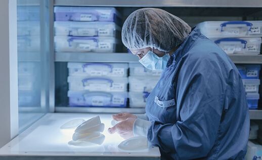 A person in medical attire examining breast implants on a lit display table, with storage boxes in the background.